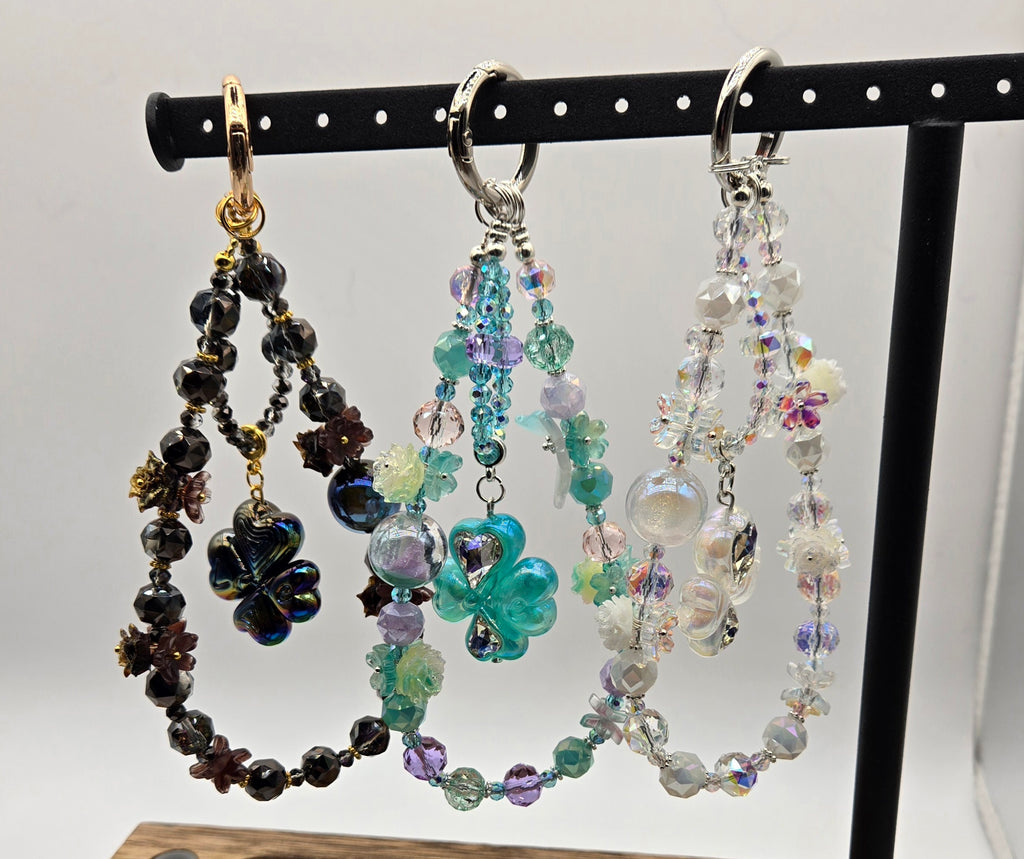 Enchanted Clover beaded phone charms in black, aqua and crystal colorways displayed on a black stand
