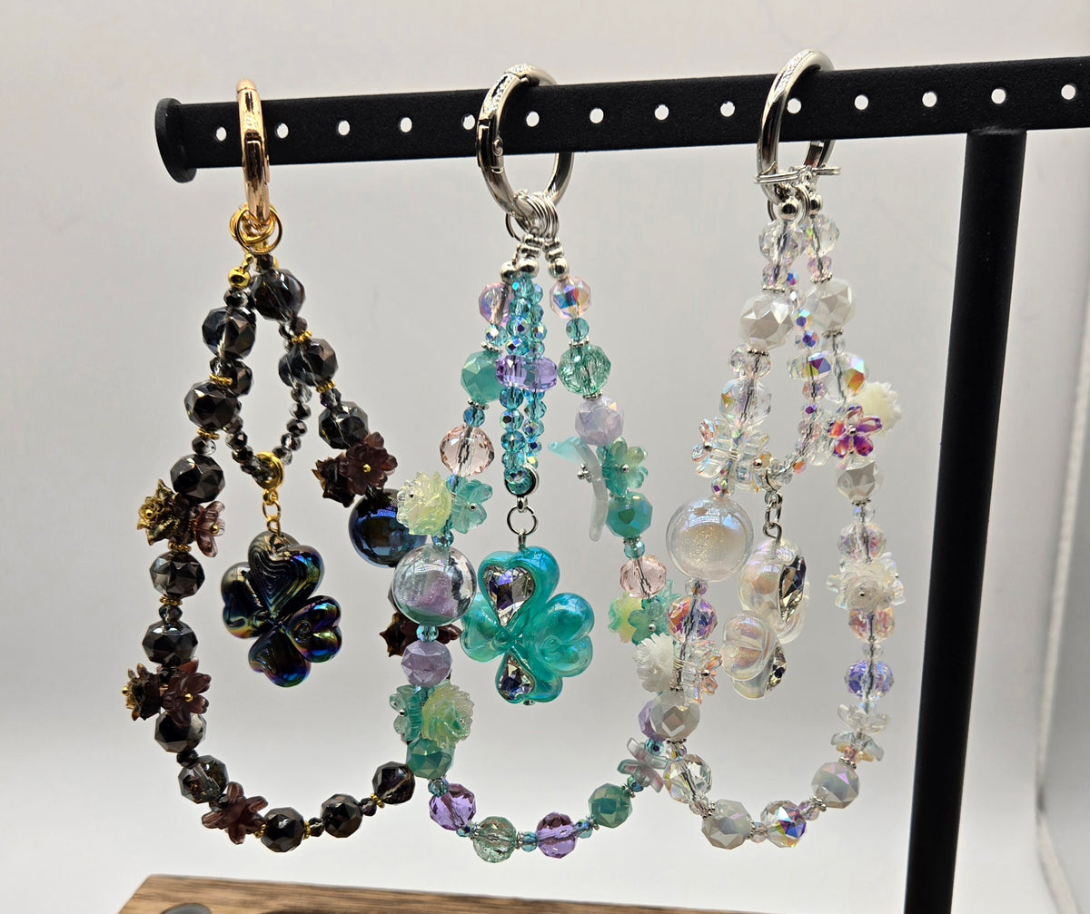 Enchanted Clover beaded phone charms in black, aqua and crystal colorways displayed on a black stand